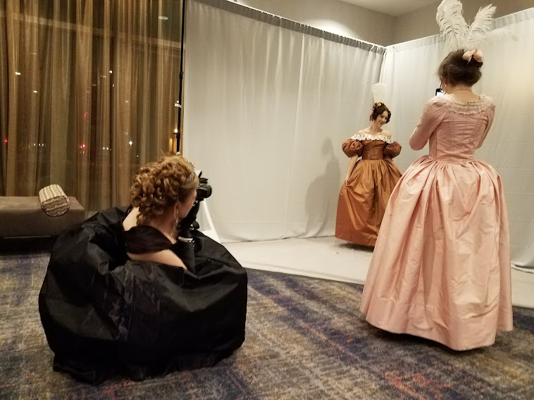 Woman in black dress kneeling with camera taking a picture of a woman in a brown dress standing in front of gray back drop while a third women in pink dress looks on.